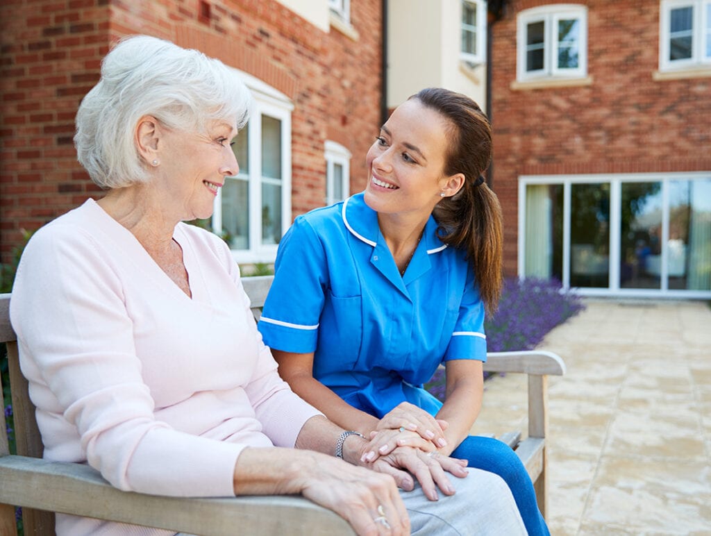 Senior Woman Sitting On Bench And Talking With Nurse In Retirement Home