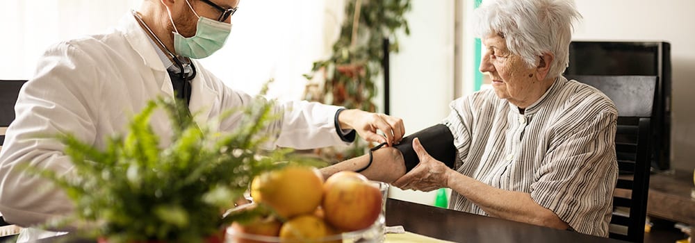 Young serious Caucasian doctor wearing face mask adjusting blood pressure gauge on senior gray-haired woman's hand during house call medical check-up