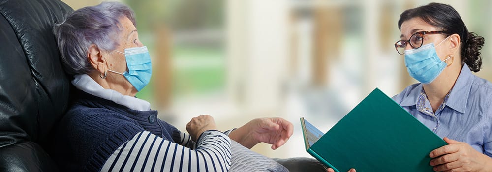 An everyday female caregiver reads books for an elderly woman. The corona-virus pandemic has made a difference. Both wearing face masks and keep their distance.